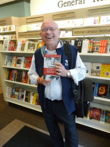 Man in blue vest, standing and holding Cleft Heart in front of bookshelves.