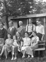 5 men standing behind their 5 wives who are sitting. The eldest woman holds a 2 y.o. female child.
