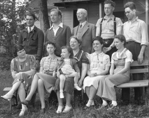 5 men standing behind their 5 wives who are sitting. The eldest woman holds a 2 y.o. female child.