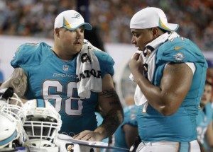 Two linemen in Dolphin turquoise jersies w/o helmets talk to one another off field