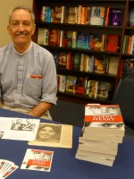 Karl Schonborn sitting at bookstore table w stack of books.