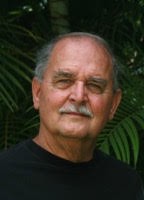 Older man with greying hair and mustache posed before palm leaves.