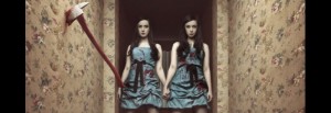 The SOSKAS twin sisters hold hands in a hallway with a bloody axe imbedded in a wall.