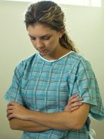 Young woman with bowed head and folded arms in hospital gown.