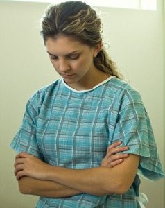 Young woman with bowed head and folded arms in hospital gown.